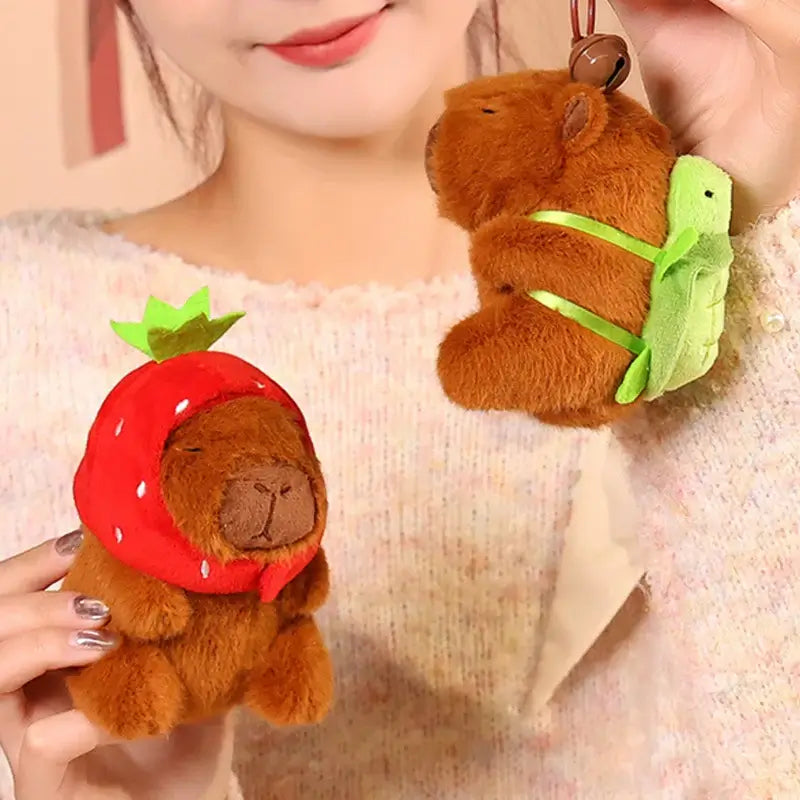 Cute brown capibara plush keychains in strawberry hat and turtle backpack, held by a person.