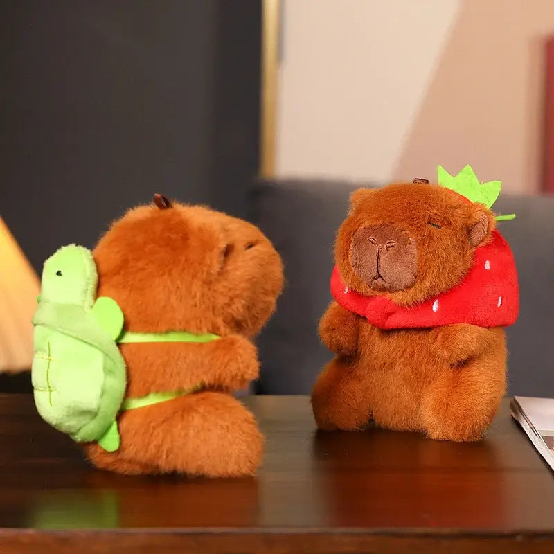 Two cute capibara plush keychains, one with a turtle backpack and the other in a strawberry hat, on a table.
