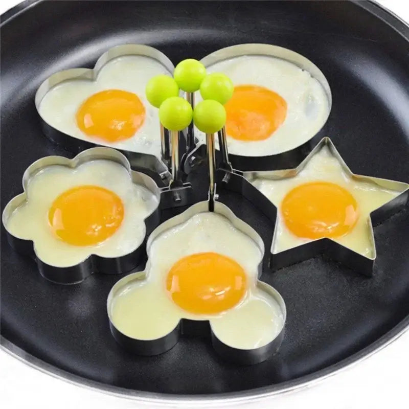 Stainless steel egg mold rings in heart, flower, star, and Mickey Mouse shapes cooking eggs in a frying pan.